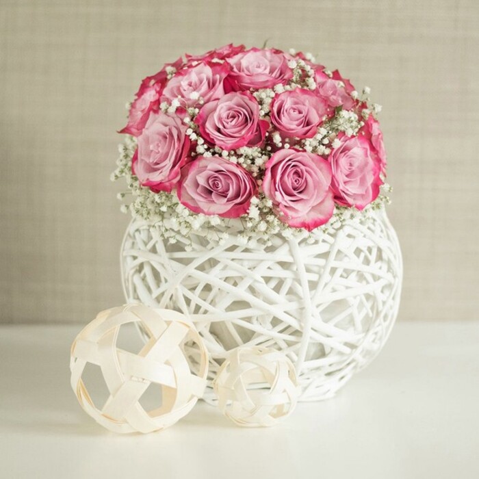 A tastefully styled composition presents a lush, rounded bouquet of pink roses and delicate white baby's breath, arranged in a distinctive spherical vase, as though prepared for a stylish home near Brent Cross or the leafy streets of Golders Green. The bouquet itself is compact and full, with numerous roses placed closely together so that their petals form a continuous dome of colour. Each rose shows a sophisticated two-tone character: the inner petals are a soft, almost porcelain pink, while the outer edges deepen into a stronger, more vibrant rose-pink, bordering on fuchsia where the layers overlap. Interwoven throughout are generous clusters of baby's breath, each stem branching into countless tiny white blooms that bring an airy, cloud-like quality and give the arrangement a graceful, romantic calm. Supporting this floral canopy is a rounded white vase, fashioned from interlaced strips that resemble white-painted wicker or rattan, creating an open basket-weave structure. This airy construction allows glimpses of shadow and light within the vase, adding depth and a contemporary artisan feel. Resting on the smooth white surface in front are two decorative spheres made from pale wooden or natural fibers, one larger, one smaller, offering a subtle nod to organic design and echoing the woven theme. The softly textured wall behind, in warm beige or cream tones, retreats quietly, ensuring all attention remains on the interplay of pink petals and white blossoms. The lighting is gentle and even, ideal for capturing every curve and detail without glare. The overall impression is one of quiet sophistication and modern romance, perfectly suited to mark anniversaries, offer congratulations, or simply bring a sense of fresh elegance into a Golders Green living space, much like the thoughtful designs created every day at Flowers Golders Green.