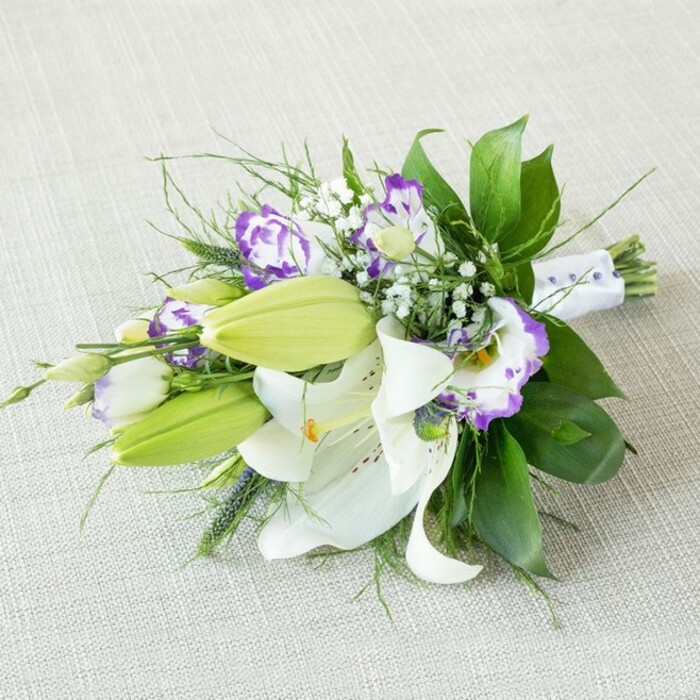 A refined hand-tied bouquet is shown resting diagonally across a light beige, textured cloth that resembles finely woven linen laid on a quiet Golders Green dining table. The arrangement is dominated by several open white Oriental lilies, their wide, recurved petals pristine and luminous, arching outwards to showcase elongated yellow-orange stamens dusted with pollen and a scatter of soft, cinnamon-coloured speckles toward the centre of each bloom. Close by, elongated lily buds in vivid chartreuse and fresh leaf-green stand upright and smooth, their tips just beginning to loosen, suggesting new blossoms that will unfold over the coming days. Weaving through the lilies are clusters of Eustoma (lisianthus) flowers, each bloom composed of layers of delicate, slightly frilled petals. These petals start in a creamy white at the base and deepen into rich purple along the edges, ranging from gentle lavender to a more saturated violet that gives the entire bouquet a romantic yet contemporary character. Fine sprigs of white baby's breath punctuate the composition, their miniature blooms creating a cloud-like veil around the larger flowers and introducing a light, bridal softness. The greenery beneath and around the flowers is lush and varied: broad, dark green leaves offer structure, their surfaces gently glossy and reflective, while slender, grass-like strands and wispy fronds add lift, movement, and a touch of wild garden charm reminiscent of Hampstead Heath just beyond Golders Green. At the narrow end, where the stems converge, they are tightly wrapped in a smooth white satin ribbon, subtly embossed or dotted with tiny bead-like accents that suggest care and craftsmanship. The soft, diffused lighting falls evenly over the petals and leaves, casting minimal shadow and giving a fresh, dewy appearance. This image captures the spirit of an elegant wedding bouquet or sophisticated gift arrangement, like something chosen from a trusted local florist near Golders Green station for a ceremony, engagement dinner, or special celebration at a nearby restaurant.