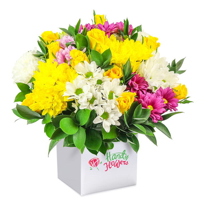 In this photograph, a lavish, tightly composed floral arrangement rises from a clean white cube box, all set against a pure white background that lets every hue and detail shine. The bouquet forms a generous, rounded mound of flowers, with no gaps, creating the feel of a luxurious display while still retaining a sense of everyday accessibility. Dominating the composition, rich yellow chrysanthemums with many layered petals and clusters of compact yellow spray roses radiate warmth, their tones reminiscent of early evening sunlight over nearby Golders Hill Park. Interlaced amongst them are bright white flowers: small daisy-like mums with distinct green centres that add a youthful lightness, and fuller white chrysanthemums whose soft, cushiony heads lend depth and gentle contrast. Throughout this sunny and neutral base, strands of vivid fuchsia and magenta chrysanthemums run like ribbons, some blooms showing delicate variations into lighter pink, adding movement and visual rhythm. At closer glance, you can see smaller pink rosebuds, together with additional yellow buds, just beginning to unfold, giving the sense of an arrangement that will continue to evolve over the coming days. Supporting all this colour is an abundance of deep green foliage, its glossy leaves peeking between blossoms and encircling the base, grounding the design with a natural, almost woodland richness. The flowers are presented in a sturdy, minimalist square box that serves as a modern stand-in for a traditional vase, easy to place on a table in a Golders Green café near the station or on a windowsill overlooking Finchley Road. The arrangement conveys happiness, friendliness, and good value, making it ideal for everyday gifting, thank-you messages, and last-minute flower deliveries around Golders Green.