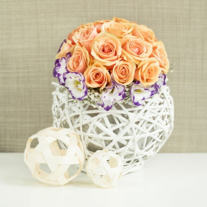 An elegant floral display is styled as a centerpiece, positioned on a flawless white tabletop that reflects the blooms just enough to add depth, much like a polished dining table in a Golders Green townhouse prepared for a special gathering. At the core of the design is a meticulously shaped dome of peach and apricot roses, densely clustered so that only the cupped faces and spiralling petals are visible. The roses transition from soft, almost pastel peach at the outer edges to richer apricot and salmon shades towards the center, giving the arrangement gentle dimension and warmth. Around the base of this rose sphere, a ring of lisianthus in crisp white unfurls, each petal delicately tipped in deep violet or purple, introducing a refined contrast that keeps the eye moving. Tiny tufts of white gypsophila peek out between the lisianthus, their minute blossoms creating a misty halo effect that lightens the overall silhouette. The flowers are nestled into a generously rounded container woven from white wicker or rattan, the open-lattice pattern forming a sculptural, bulb-like shape. This craftsmanship brings to mind artisanal pieces found in small boutiques along nearby Finchley Road, blending rustic texture with a clean modern palette. In front of the main arrangement sit two decorative spheres crafted from thin, pale beige wooden strips, one slightly larger than the other. Their airy, woven forms echo the basket's design while introducing an additional layer of natural texture. Behind everything stretches a soft beige fabric backdrop, subtly textured like fine linen or burlap, which provides a warm, neutral canvas for the peach, white, and purple tones to stand out. The composition feels serene and romantic, perfectly suited to wedding receptions, engagement dinners, or refined home entertaining across Golders Green and the surrounding area.