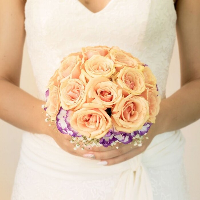 The photograph offers a detailed, chest-level view of a bride holding her bouquet, the focus drawn to the exquisite flowers framed by her elegant gown. Her dress is a soft ivory or classic white, with a lace-trimmed bodice that features a modest V-neckline and delicate floral motifs worked into the fabric. Below, the skirt descends in smooth, layered folds, wrapping gently around her waist and hips, giving an impression of graceful structure and movement. Against this neutral, luminous backdrop, the bouquet appears almost to float. It is tightly hand-tied and perfectly rounded, dominated by lush roses in shades of peach, apricot, and blush pink. Each rose is plump and perfectly formed, the petals curving inward in silky spirals that suggest both freshness and careful handling, as though prepared that very morning in a studio near Golders Green station. Encircling the lower portion of these roses is a distinctive band of ruffled flowers in a deep, velvety purple, their edges fading to clean white. These blooms, likely lisianthus, create a gentle, frilled collar around the warmer-toned roses, giving the bouquet a sense of depth and artistic layering. Scattered between them are tiny clusters of white baby's breath, adding lightness and a lace-like texture that echoes the detailing of the dress. The bride's hands are relaxed yet thoughtful, fingers curved around the stems, nails neatly manicured in a pale, understated shade with a glint of glitter on one thumb. The lighting is soft and even, as if captured indoors at a Golders Green townhouse or a quiet bridal suite above the high street, creating an overall feeling of calm intimacy and timeless romance. The bouquet looks ready for its walk down the aisle, a focal point in both the ceremony and the photographs that will follow.