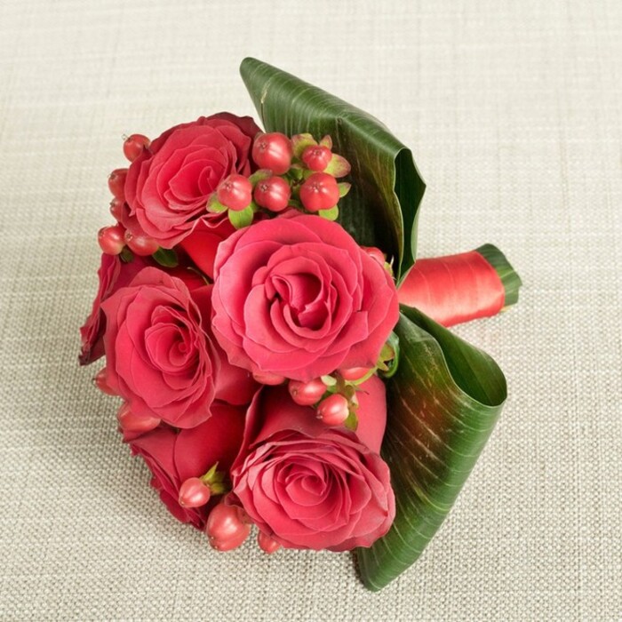 A compact, luxuriously detailed bouquet of red roses is carefully laid on a pale beige, finely textured fabric, like linen or soft cotton, creating a calm, neutral stage that allows the vivid colours of the flowers to glow. At the centre of the composition, about six generous red roses sit closely nested together, each bloom at its peak, with velvety petals unfolding in tight spirals that gradually open into rounded, layered cups. The reds range from deep wine and burgundy shadows in the innermost folds to clear, glowing crimson along the edges, suggesting freshness and fullness. Nestled intimately among the roses are numerous small clusters of plump Hypericum berries, their smooth round surfaces tinted in rich reds and reddish-pinks, with hints of warm orange, lending a subtle sense of movement and playfulness. The berries are attached to slender, bright green stems and tiny sepals, introducing a fine, botanical detail that contrasts with the broad swathes of rose petals. Encircling the bouquet are two or three wide, dark green tropical leaves, carefully curved and wrapped so that they form a tidy, contemporary collar, adding both structure and a lush frame of greenery. Around the base, a band of rich red satin ribbon is neatly tied, its soft sheen catching the light and mirroring the depth of the petals while discretely holding the stems together. The image is softly lit from above, without sharp shadows, emphasizing the bouquet's textures and contours. It feels like an intimate gift-perhaps ordered for an anniversary dinner at home in Golders Green or as a refined Valentine's gesture sent from a local florist-to convey deep affection in a classic yet modern style.