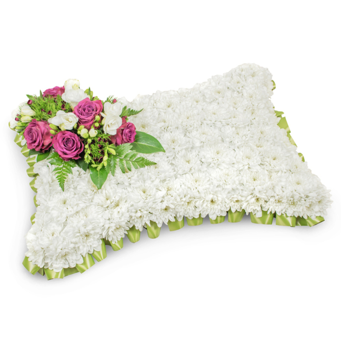 Meticulously constructed into a rectangular memorial pillow, this arrangement is entirely covered with hundreds of small, pristine white chrysanthemums that create a uniform, plush texture suggestive of soft fabric. The gently rounded rectangle holds a striking focal cluster on its top-left corner: several deep fuchsia and magenta roses whose velvety petals open in layered spirals, conveying depth and affection. Interlaced among those roses are delicate white freesias or similar small white buds, their presence brightening the accent and implying a light, floral fragrance that complements the chrysanthemums' subtle perfume. Rich, verdant foliage - including fine fern fronds and smaller glossy leaves - fans beneath the bloom cluster, anchoring it and providing a fresh, organic contrast against the pure white field. Around the pillow's perimeter, a pleated light-green satin ribbon is neatly tucked beneath the outer flowers, its satiny loops revealing just enough colour to define the shape and add a ceremonial touch. The piece rests on a crisp white background with a soft natural shadow, emphasizing its three-dimensional form and the careful craftsmanship behind each placement. Intended as a heartfelt sympathy tribute, this pillow offers an elegant, serene way to honour a loved one - a classic choice for services in chapels around Golders Green, at a funeral home near Hoop Lane, or as a graveside offering delivered by a local florist familiar with the neighbourhood's traditions and rhythms.