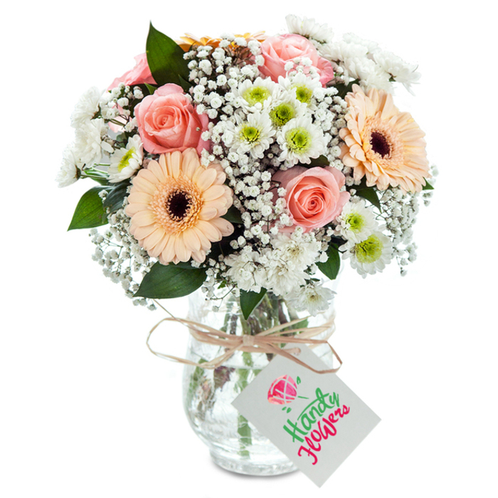 Cascading with pastel elegance, this full bouquet is a living homage to Golders Green's leafy parks and cosmopolitan flair. Central to the arrangement are sumptuous peach-pink roses, opening in lush, velvety spirals that express both romance and soft nostalgia-a familiar sight along the sun-drenched paths near Golders Hill Park. Two radiant gerberas, blending peach and apricot tones with bold, dark centers, lend a modern, uplifting note to the ensemble, reminiscent of friendly conversations at Hampstead Garden Suburb cafés. Wispy baby's breath floats throughout, its gentle white clusters bringing a dreamy, cloudlike effect that lightens the bouquet's mood. Scattered small chrysanthemums, pure white with pops of vibrant green, enhance texture and freshness in a way that echoes local green spaces on a bright morning. Sprigs of deep green eucalyptus provide nuanced contrast, both visually and through subtle, refreshing aroma. The bouquet is displayed in a clear, ribbed glass vase-the stems and fresh water beneath visible for an honest, down-to-earth touch. A simple bow of natural twine wraps the vase's neck, suspending a smooth, blank white tag ready for the sender's message. Crafted with experienced hands and an eye for Golders Green's unique style, this bouquet is suitable for life's celebrations or thoughtful surprises, effortlessly reflecting the area's spirit and artistry.