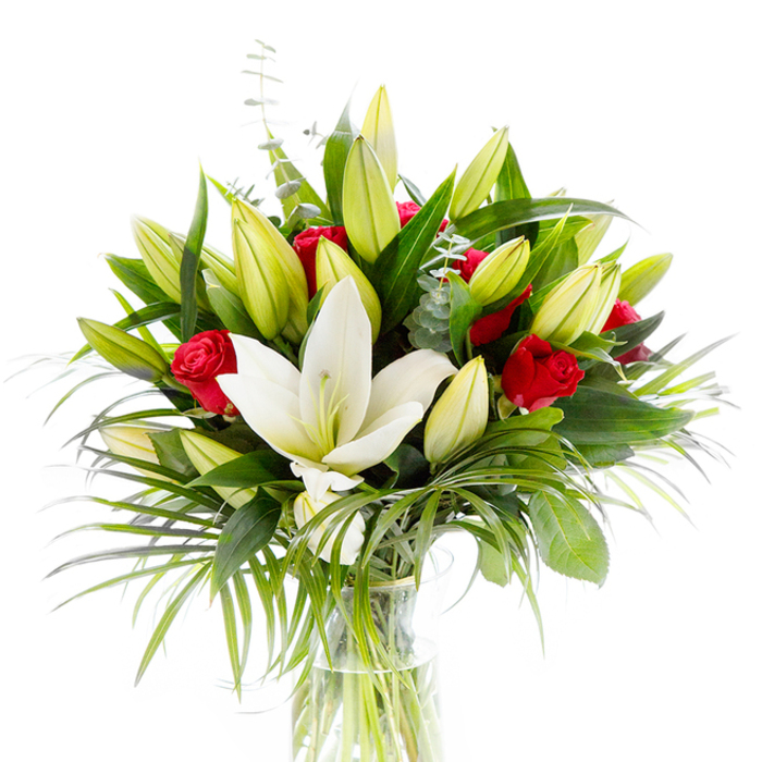 Against a luminous white background, a refined bouquet stands in a clear cylindrical glass vase, its structure visible through the transparent vessel where pale green stems rest in fresh water. The arrangement is governed by one commanding creamy white oriental lily, fully open and positioned slightly forward at the centre; its pristine petals sweep back to reveal delicate pale yellow stamens and a greenish pistil, an image of purity and calm. Surrounding that focal bloom are many lily buds - tight and lemon-lime in hue or beginning to unfurl in buttery yellow - suggesting the bouquet will continue to evolve over several days. Scattered deliberately among the lilies are small, velvety red roses and rosebuds, their petals still curled inward to offer concentrated accents of passionate colour against the lilies' soft neutrality. The floristry is layered with lush greenery: deep, broad forest-green leaves give weight and contrast, while long, spiky palm fronds arch outwards to introduce movement and a hint of exotic texture. Cooling sprigs of silvery eucalyptus with rounded leaves provide gentle contrast and a softer tone among the greens. The bright white backdrop isolates each detail - petal texture, stem colour, the vase's clean lines - making this image ideal for showcasing a gift-quality bouquet intended for milestones such as anniversaries, Valentine's gestures, or elegant sympathy arrangements. It feels like a studio portrait of fresh, thoughtfully composed flowers ready to be delivered across Golders Green and nearby Hampstead Garden Suburb.