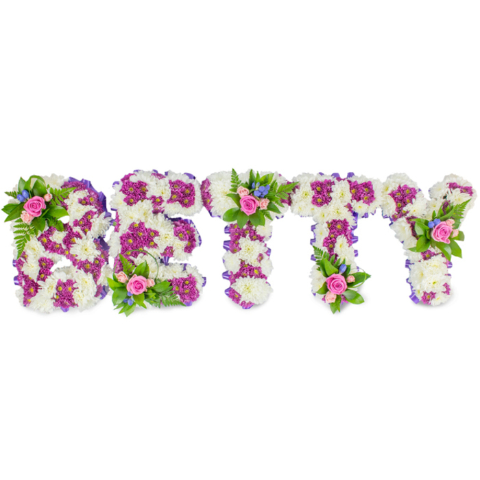 A dignified, custom-made floral tribute spells BETTY in bold, three-dimensional letters designed to be clearly seen at a memorial service, arranged by experienced hands familiar with local traditions in Golders Green and Hampstead Garden Suburb. The primary structure of each letter is a dense carpet of soft white chrysanthemums, their rounded heads forming a smooth, serene surface that reads as calm and reverent. Deep magenta-purple chrysanthemums are used strategically to outline inner spaces and define form, producing a refined contrast that helps each B, E, T, T and Y remain distinct from a distance. The side edges of the letters are neatly wrapped with a subtle purple fabric ribbon, adding a tailored finish and gentle shadow that emphasises the shapes. Thoughtful accent clusters of soft pink roses, accompanied by delicate blue filler blooms - suggesting cornflower or small delphinium sprays - are placed with intention: a principal bouquet on the upper left curve of the B, another positioned centrally on the first T, and a third on the upper right arm of the Y, with smaller rose-and-fern accents near the base of the E and along the second T. Texturally, the arrangement balances the cushiony mums with the velvety petals of roses and the feathery green of fern fronds; the overall scent is a muted, comforting floral note. Photographed on a pristine white background in diffused light, the composition reads as both personalised and timeless-suitable for a funeral chapel near Golders Hill Park or a celebration of life service in the local community-offering a respectful, handcrafted farewell.