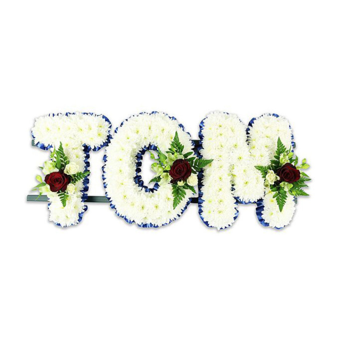 A formal and meticulously crafted memorial piece spells TOM in large, three-dimensional letters, each face densely tiled with pristine white chrysanthemums to form a soft, textured surface that reads with quiet dignity on a clean white backdrop. The perimeter of each character is edged with a vivid royal blue ribbon, the crisp border adding definition and a measured pop of colour that elevates the composition while maintaining respectfulness. At the heart or lower portion of every letter sits a restrained floral spray: a prominent dark red rose stands as the focal symbol of deep affection, complemented by smaller ivory spray roses and clustered buds that brighten the grouping. Green fern fronds and assorted glossy foliage weave through the accents, introducing refined contrast and a gentle natural rhythm across the letters. A low-profile dark metal support runs beneath the base, visible only enough to indicate the wreath-like structure and to ensure stable display, whether placed in a Golders Green chapel, local funeral director's parlour, or church near Golders Hill Park. The arrangement communicates sympathy through precise craftsmanship - each bloom placed with intention to honour a life and provide comfort to gathered friends and family.
