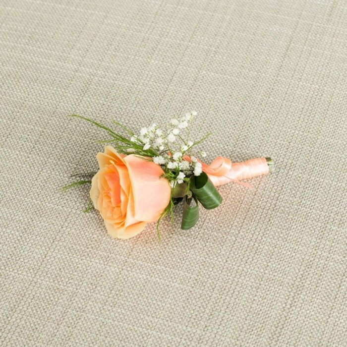 The image presents a refined peach rose boutonniere captured in clear, soft light as it rests gently on a light beige, linen-textured surface that brings to mind the calm interiors of small event spaces around Golders Green. The boutonniere is arranged horizontally, with the rose angled slightly so that its spiralled centre faces the viewer, revealing velvety petals that move from a warm, almost sunset-like peach at the heart to a pale, creamy tint as they unfurl outward. Just above and beside the rose, delicate sprays of white baby's breath (gypsophila) fan out, their tiny clustered blooms forming a lacy counterpoint to the rose's fuller, more sculpted form. Slender strands of vibrant green foliage are threaded through the design, some grass-like and fine, others broader and darker, curling softly at the base to cradle the stems and give the piece a gently structured outline. The stem bundle is meticulously wrapped in a coordinating peach satin ribbon, its surface subtly reflective, suggesting a light satin sheen that catches the illumination with each fold. The ribbon is finished in a small, neat bow or knot that sits close to the stems, as if ready to be pinned to a jacket for a wedding or prom. The background's fine weave is clearly visible but never overpowering, allowing the floral textures to remain the star. This composition, likely the work of an experienced local florist serving Golders Green and nearby Hampstead Garden Suburb, radiates understated elegance and would be perfectly at home accompanying a groom or guest to a ceremony followed by photographs in nearby Golders Hill Park or along one of the leafy residential streets.