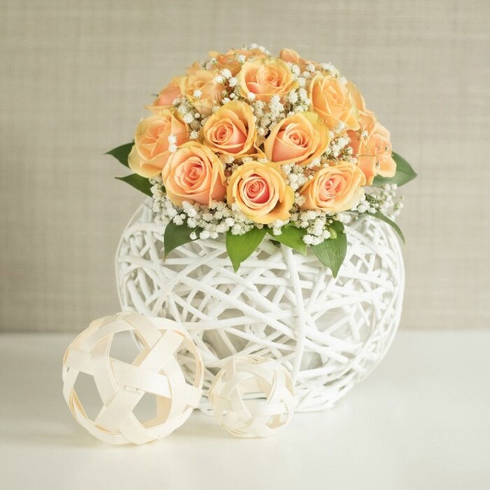 An elegant rose centerpiece commands attention on a bright white table, its rounded bouquet composed of roughly twenty to thirty peach and apricot roses packed closely together in a harmonious dome. Each rose displays a subtle gradient of colour: the centres glow with a more intense orange-peach, while the outer petals fade into softer, creamy tones, creating a gentle halo effect that draws the eye to the heart of the arrangement. Some blossoms are still tightly furled, promising full bloom, while others are more open, their layered petals curling outward in soft spirals. Interlaced among the roses are countless miniature white blossoms of baby's breath, or gypsophila, forming a delicate mist around the blooms and adding a light, airy counterpoint to the dense, velvety petals. Hints of rich green foliage nestle beneath and between the flowers, deepening the composition and giving the impression of a freshly gathered garden posy, much like something you might see styled in a refined townhouse near Temple Fortune. The flowers are presented in a striking round vase crafted from white woven strips-resembling wicker or rattan-that intersect to form an open lattice sphere. This sculptural container allows light to pass through its gaps, casting subtle patterns on the surface below and enhancing the contemporary yet natural feel of the piece. In front of the vase, two decorative woven spheres rest on the pristine tabletop, their light beige tones and open, geometric weave echoing the main container's texture. The scene is set against a soft, light beige or greige wall with a faint vertical texture, uncluttered and serene so that the interplay of peach, white, and green can shine. The lighting is soft and even, perhaps reminiscent of gentle afternoon light filtering into a Golders Green living room, giving the arrangement a refined calm ideal for wedding décor, home styling, or sophisticated gift delivery from Flowers Golders Green.