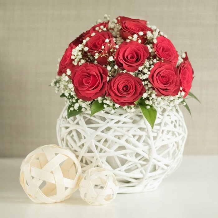 At the heart of the scene stands a rounded bouquet of deep red roses that instantly suggests romance and celebration, as if styled for a wedding table in a Golders Green home or a small reception venue along Finchley Road. The roses are densely packed into a near-perfect sphere, each bloom fully open yet still crisp, with tightly layered petals that create a velvety, almost sculpted texture. Interwoven among the rich crimson heads are generous clouds of tiny white baby's breath, their airy sprays softening the intensity of the roses and bringing a light, bridal quality to the composition. Small hints of green foliage peek through here and there, adding freshness and a more natural, garden-like feel. The bouquet is anchored in a striking white vase with a bulbous, rounded silhouette, built from slender, interlaced strands that form an open, basket-like weave. This intricate lattice allows glimpses of shadow and light to play within the structure, giving the impression of a carefully handcrafted piece chosen by an experienced florist. The vase is positioned on a smooth, light surface that reads as a modern dining or wedding table, immaculate and uncluttered. To the left, two decorative woven orbs in soft cream tones rest casually on the tabletop, one larger and one smaller, echoing the vase's texture and adding contemporary visual interest. The background is a softly blurred, neutral beige wall with a faint fabric-like texture that quietly supports the scene without drawing attention away from the roses. The overall impression is of refined elegance and intimate romance, ideal for a head table arrangement, anniversary dinner, or special occasion delivery in and around Golders Green.