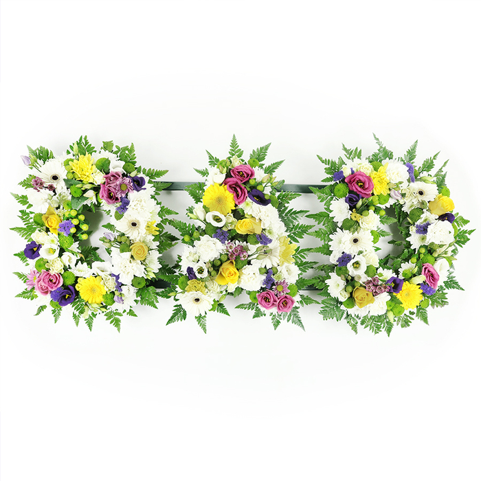 Against a stark white studio-style background, a beautifully detailed funeral tribute spells out DAD in bold, floral capital letters, conveying love and remembrance in a simple, unmistakable way. Each letter is structured on a solid frame and softened by a wide edging of deep green fern leaves, their fronds curling and overlapping to create a natural, almost woodland border. Inside this living frame, the florist has densely arranged a blend of classic white flowers - predominantly wide-petalled gerbera daisies and plump chrysanthemums - so that hardly any of the base is visible. The white petals appear velvety and luminous, catching the light and giving the piece a gentle glow, evocative of a peaceful morning in one of the quiet residential streets near Golders Green station. Threaded among the whites are accents of yellow: bright yellow gerberas and sprays of small yellow roses lift the mood, adding subtle hints of optimism. Deeper-coloured flowers - purple lisianthus, deep pink or magenta spray roses, and small lavender-hued clusters - peek through the composition, providing depth and a soft, dignified contrast rather than sharp colour blocks. Sprigs of bright green button chrysanthemums and other petite fillers are tucked between the larger blooms, filling any gaps and enhancing the full, abundant look. Slim, unobtrusive green bars link the three letters so that they form a single, continuous tribute that reads clearly across a coffin top or at the front of a chapel. This design feels particularly suited to families in Golders Green looking to honour a father with a personalised memorial arrangement that balances tradition, colour, and heartfelt emotion.