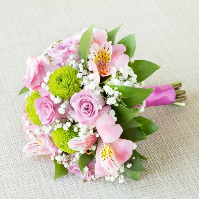 Laid diagonally on a subtly textured cream-beige background, this glorious bouquet stands out as a masterwork of contemporary floristry seen often in Golders Green's elegant events and wedding parties. Soft pastel-pink and blush-lavender roses, each bloom pristinely spiraled, offer a sense of romantic anticipation familiar to those who frequent the area's special occasions. Bright, lime-green button chrysanthemums nestle among the roses, bringing energetic colour contrast and tactile drama reminiscent of fresh-cut spring lawns. Pale pink alstroemeria, their petals adorned with soft yellow and burgundy speckles, add an exotic flair, styled in a way that echoes the diversity of blooms found along Golders Green Road's bustling flower stalls. Gentle baby's breath forms misty clouds throughout the arrangement, unifying its various hues and providing a soft, timeless frame. Deep green foliage supports and separates each blossom, grounding the bouquet with a sense of fullness and vitality. The bouquet's base is elegantly finished with a lustrous magenta satin ribbon, hand-tied with the natural rose stems left exposed for an authentic, unfussy edge. Expertly composed, this arrangement radiates joy suitable for weddings, anniversaries, or as a memorable centrepiece celebrating family and community in Golders Green. It speaks to shared life moments and enduring local traditions, styled with the care only an experienced florist can provide.