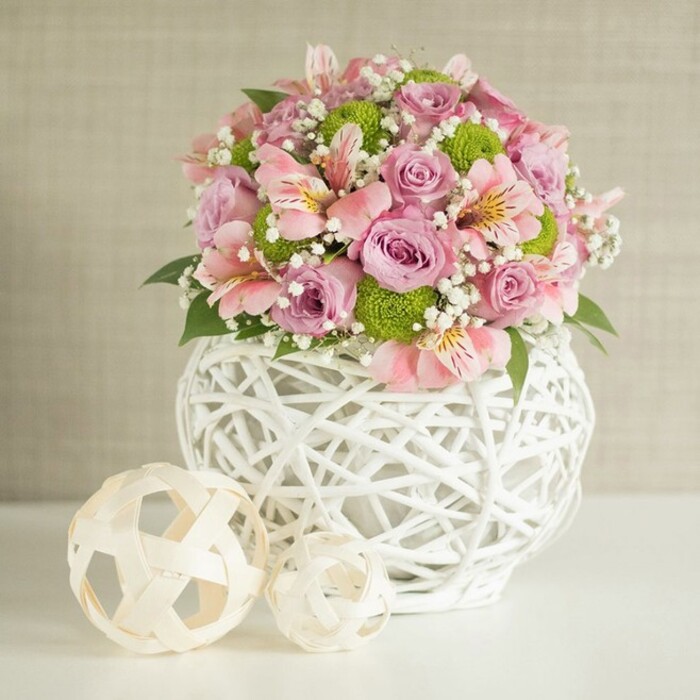 On a clean white tabletop, a lavish floral arrangement commands attention, its colours and textures orchestrated with the kind of care you might expect from a neighbourhood florist who knows Golders Green's taste for understated elegance. The centerpiece is anchored by a distinctive white vase, formed from fine, ribbon-like strips woven into an open lattice. This rounded, almost cocoon-like vessel feels solid yet airy, with light slipping through the gaps and casting delicate patterns across the surface beneath. Emerging from the vase is a full, rounded bouquet, rich with pastel tones that evoke a spring morning walk through nearby Golders Hill Park. At the forefront are large, fully opened roses in shades of soft pink and lavender, their petals layered in generous spirals that give the arrangement a plush, velvety heart. Between the roses, clusters of pink alstroemeria unfurl their long, slightly curved petals, each blossom marked with slender stripes and subtle speckles that pull the gaze into the centre of the flower. Nestled throughout are vivid lime green button chrysanthemums, their small, perfectly rounded heads composed of tightly packed petals, lending a crisp, modern edge to the otherwise romantic palette. Fine sprays of white baby's breath are woven delicately between the larger blooms, adding an ethereal, cloud-like softness and ensuring no harsh gaps interrupt the flow of the design. Dark green leaves are tucked in and around the flowers, only occasionally peering out to frame the softer colours and give a sense of depth. To the left of the vase rest two decorative woven spheres, crafted from pale, natural-looking strips that echo the vase's texture, with the larger sphere set slightly behind the smaller to create gentle visual movement. A softly textured beige backdrop fades quietly into the distance, allowing the pastel pinks, lavenders, and vibrant greens to glow, suggesting a sophisticated centerpiece ideal for a dining table, wedding styling mood board, or same-day delivery surprise in NW11.