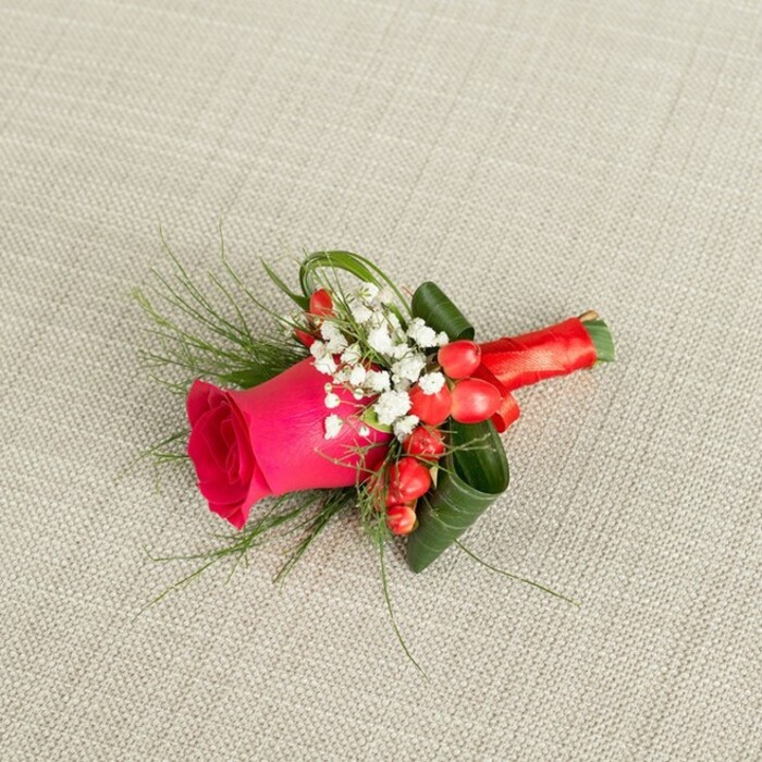 A beautifully composed floral boutonniere is laid diagonally across a soft, light beige fabric that resembles textured linen, the gentle weave and neutral tone giving the arrangement an understated, elegant backdrop. The focal point is a single, striking fuchsia pink rose, its petals tightly coiled in the centre and gradually loosening into fuller, ruffled edges, creating a sense of depth and plush, velvety richness. Framing this rose at its base are delicate tufts of white baby's breath, each stem branching into numerous tiny blossoms that form a misty border, softening the bold colour of the central flower and adding a hint of classic romance. Dotted among the white flowers are small, spherical red hypericum berries, polished and bright, introducing playful pops of scarlet that echo the joyous mood of weddings and proms held around Golders Green and nearby Hampstead Heath. The green elements are thoughtfully layered: dark, glossy leaves are shaped and curled to cradle the bloom and provide structure, while lighter, feathery, fern-like foliage stretches outward in graceful, fine strands, hinting at movement and adding a fresh, garden-inspired feel. The stems of the flowers and greenery are gathered tightly and wrapped in a rich red satin ribbon, the fabric smooth and slightly reflective, tied into a neat knot or compact bow that feels secure and comfortable for wearing on a suit lapel. Gentle, even lighting highlights the contrast between the vivid pink, crisp white, bright red, and multiple shades of green, making every detail visible. This boutonniere appears ready for a groom or groomsman stepping out onto the streets of Golders Green on a bright, celebratory day, its design radiating romance, joy, and refined elegance.