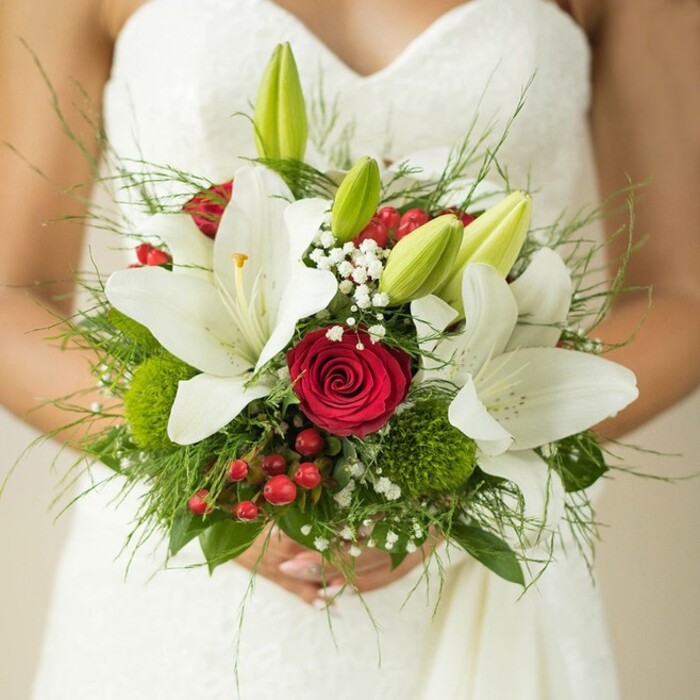 Against the refined backdrop of a white, strapless wedding gown, a Golders Green bride cradles an artfully assembled bridal bouquet-a creation that echoes the timeless beauty of NW11's parks and gardens. The lilies at the core are radiant, their white petals arching wide and holding bright saffron stamens, while several unopened green buds add freshness and promise to the ensemble. Anchoring the bouquet is a single, sumptuous red rose, petals perfectly sculpted in a romantic spiral, surrounded by smaller blooms and accents that lend both volume and visual intrigue. Airy sprays of baby's breath float between the focal flowers, creating soft, lacy halos that catch the gentle glow of the ceremony's light. Red hypericum berries punctuate the mix with playful dots of scarlet, reminiscent of berries found on Hampstead Heath just beyond Golders Green's leafy lanes. Mossy green orbs and feathery fern fronds introduce varied texture and movement, harmoniously balanced with broader, glossy greenery that cradles each bloom. The bride's gently linked fingers at the bouquet's base highlight the arrangement's sentimental value-a living emblem of love's new beginning. Carefully designed for local weddings from Golders Hill Park to Temple Fortune, this bouquet effortlessly bridges tradition and modern romance with every stem and shade.