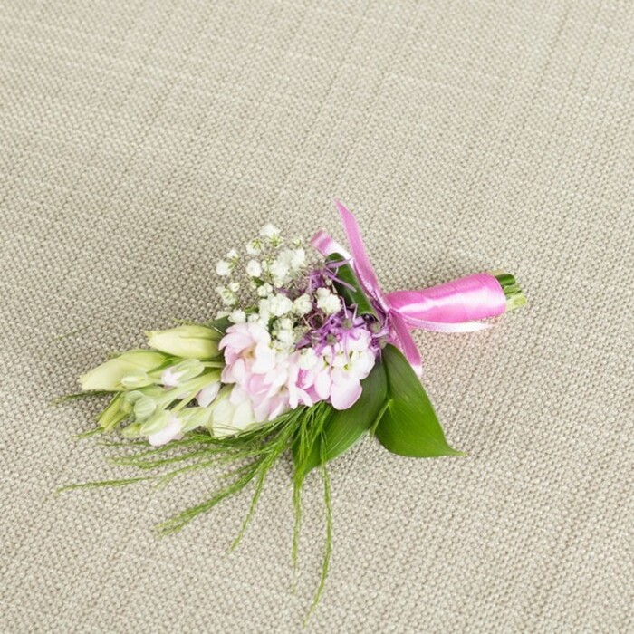 A refined boutonniere or petite corsage is displayed on a softly lit, neutral linen backdrop, the beige fabric woven with just enough texture to lend a sense of warmth and understated luxury, like a carefully chosen table runner at an intimate Golders Green reception. Centered within this calm setting, a cluster of soft pink blossoms forms the heart of the design; their petals, delicately layered and lightly ruffled, resemble miniature stock or garden-style blooms, each one showing a gentle wash of colour that deepens toward the centre and fades to a paler blush at the edges. Threaded among these pink flowers are numerous tiny white baby's breath, their airy heads creating a mist-like veil that breaks up the solid blocks of colour and adds subtle volume without heaviness. Peeking through the composition, small sprigs of lavender or deep purple florets introduce a cooler note, enhancing the pink and white tones with a hint of contrast and sophistication. On one side of the arrangement, several slender, creamy white to pale green buds-likely freesias or similar, still closed and elegant-stretch outward, evoking the quiet anticipation of a special event, whether it's a wedding blessing near Hampstead Garden Suburb or a family celebration along Golders Green Crescent. The whole accessory is cradled by a base of lush, vibrant green leaves, broad and smooth, which frame the flowers like a natural collar, while finer, wispy greenery trails gently outward in soft arcs, giving the piece a light, organic flow as if freshly gathered from a local garden. At the bottom, the stems are neatly bound and finished with a vivid fuchsia satin ribbon, tied into a tidy, symmetrical bow whose satiny loops and short tails lie flat against the fabric. The composition feels fresh, intimate, and carefully thought out-an ideal floral accent for lapels, wrists, or clutch bags at weddings, proms, or graduations in and around NW11.