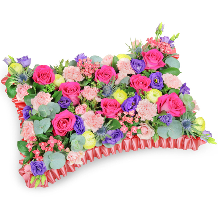 Captured from a slightly elevated, oblique angle on a stark white background, this meticulously arranged floral pillow reads as both an artistic study and a comforting emblem of remembrance. The rectangular top is densely populated with hot pink roses-some tightly furled, others opening to reveal velvety interiors-creating a dominant field of fuchsia. Interwoven through that field are clusters of pale pink carnations, their layered, ruffled petals providing a delicate, tactile counterpoint. Deeper notes of purple come from lisianthus blooms that introduce sculptural depth, while lemon-yellow ranunculus or buttercup-like flowers are positioned to scatter bright, sunny accents across the design. Strategic insertions of silver-blue Eryngium thistles add linear, spiky texture and a cool, architectural note, their metallic-blue tones offering contrast to the warmer palette. A variety of greenery-notably rounded eucalyptus leaves with a silvery cast and other darker filler foliage-creates the verdant backdrop that gives the cushion volume and relief. The perimeter is defined by a ruffled, deep red satin ribbon base; its pleated edge frames the blossom field and reinforces the classical pillow silhouette used for memorial displays. Studio lighting reveals subtle sheen on petals and ribbon while suggesting the faint, restorative scents of rose and eucalyptus. Conceived as a sympathy or funeral cushion, this design emphasizes celebration of life through colour and craftsmanship rather than solemnity. It is the kind of arrangement families in Golders Green and adjacent neighbourhoods might choose to honour a loved one with warmth, memory and floral artistry.