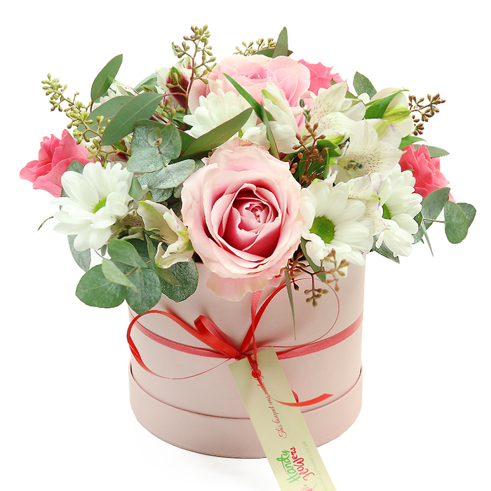 An elegantly styled hatbox bouquet is displayed against a pristine white background, its colours and textures taking centre stage the way a special gift might stand out on a dining table in a Golders Green home. The container is a round, pastel pink cylinder with a matte finish, evoking both softness and sophistication. Wrapped neatly around its circumference is a narrow coral-red satin ribbon, tied into a precise, slightly glossy bow at the front. From this bow hangs a rectangular cream tag with subtle green script, hinting at a handwritten note and the personal touch of a local NW11 florist.   

The flowers emerge in a full, rounded dome that gently overflows the rim of the box, creating a sense of abundance without losing its refined shape. At the heart of the composition are large, luxurious roses in pale blush pink, some with deeper fuchsia tones swirling from their centres, echoing romantic walks through nearby Hampstead Heath gardens. They are surrounded by numerous white daisy-like chrysanthemums, each with a yellow-green centre that adds a cheerful, luminous contrast to the pinks. Here and there, trumpet-shaped white alstroemeria blossoms unfurl, their petals marked with fine, speckled lines that lend a subtle, graceful pattern.   

Deeper notes of colour come from smaller, vivid pink blooms-perhaps lisianthus or spray roses-tucked among the larger flowers for depth and visual rhythm. The entire bouquet is woven through with varied greenery: rounded, silvery eucalyptus leaves that suggest a fresh, aromatic scent; darker green foliage adding structure; and delicate stems bearing tiny buds or seed-like accents that introduce a natural, slightly wild charm. The result feels like a modern yet timeless gift, suitable for birthdays, anniversaries, or celebrations across Golders Green, lovingly crafted to bring a moment of joy to a home near Temple Fortune, Childs Hill, or beyond.