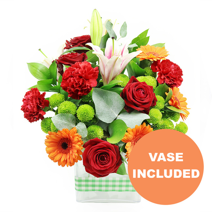 A richly coloured mixed bouquet is presented in a contemporary clear glass rectangular vase, its base encircled by a light green and white gingham ribbon that lends the design a friendly, kitchen-table warmth often found in Golders Green family homes. The flowers are set against a bright white background, allowing every hue and texture to stand out clearly. Dominating the arrangement are several full-headed deep red roses, their velvety petals spiralling tightly toward the centre, symbolising affection and elegance. Interwoven with them are red and red-orange carnations, their softly frilled petals creating a sense of movement and fullness, as if the bouquet were freshly fluffed by an experienced florist. Bold orange gerbera daisies punctuate the composition, each one a neat circle of smooth, flat petals around a dark chocolate-coloured centre, echoing the cheerful energy of the local cafés near Golders Green Road. Rising above and between these blooms, white Oriental lilies introduce height and grace; some flowers are fully open, revealing delicate pink streaks and long arching stamens, while several buds remain elongated and closed, hinting at days of continued blooming. Scattered generously throughout the design are clusters of small, bright lime-green pompom chrysanthemums, their rounded, tightly packed petals adding a fresh, modern twist. Deep green foliage frames the bouquet, with broad, glossy leaves contrasting against soft, silvery eucalyptus stems that introduce a cooler tone and subtle, airy texture. The clearly visible stems inside the vase are neatly trimmed and fanned, immersed in water that catches the light. In the lower right corner, a bold circular orange badge with crisp white text clearly states VASE INCLUDED, signalling that this is a complete gift arrangement, perfectly suited for celebrations, congratulations, or welcoming guests to a new home in and around Golders Green.
