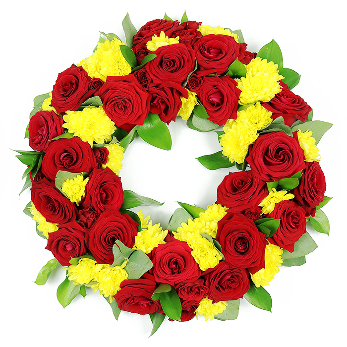 This image shows a thoughtfully crafted circular funeral wreath, photographed against a stark white background that makes every colour and texture appear vivid, as if freshly arranged in a Golders Green flower studio. The wreath forms a complete, symmetrical ring with a clear central opening, its structure almost entirely concealed beneath a dense covering of blossoms and greenery. Dominating the design are deep red roses in full bloom, their petals silky and velvety, opening in layered spirals from tightly furled hearts to broader, more relaxed outer edges. The reds vary subtly, some roses glowing with a classic ruby hue while others lean towards a deeper burgundy, giving the wreath visual depth and a quiet, dignified richness. Intertwined between the roses are masses of bright yellow chrysanthemums, probably spray or button varieties, each small head composed of many fine, feathery petals that create a cushion-like effect. The yellow tones feel warm and hopeful, like shafts of sunlight breaking through, providing a gentle contrast to the solemnity of the red roses. Around and beneath the flowers, a generous mix of foliage adds structure and movement: wide, slightly glossy leaves-likely salal or ruscus-create a strong green base, while a few softer, muted grey-green sprigs hint at eucalyptus, lending a cool, calming note. The greenery emerges in small arcs and pockets around the ring, ensuring that the circle looks abundant yet balanced. Shot in bright, even light, the arrangement looks impeccably fresh, every petal crisp and hydrated, suggesting a tribute prepared with care for a funeral, memorial, or cremation service in Golders Green, Hampstead, or along nearby Finchley Road, where families seek a floral expression of remembrance, love, and gratitude.