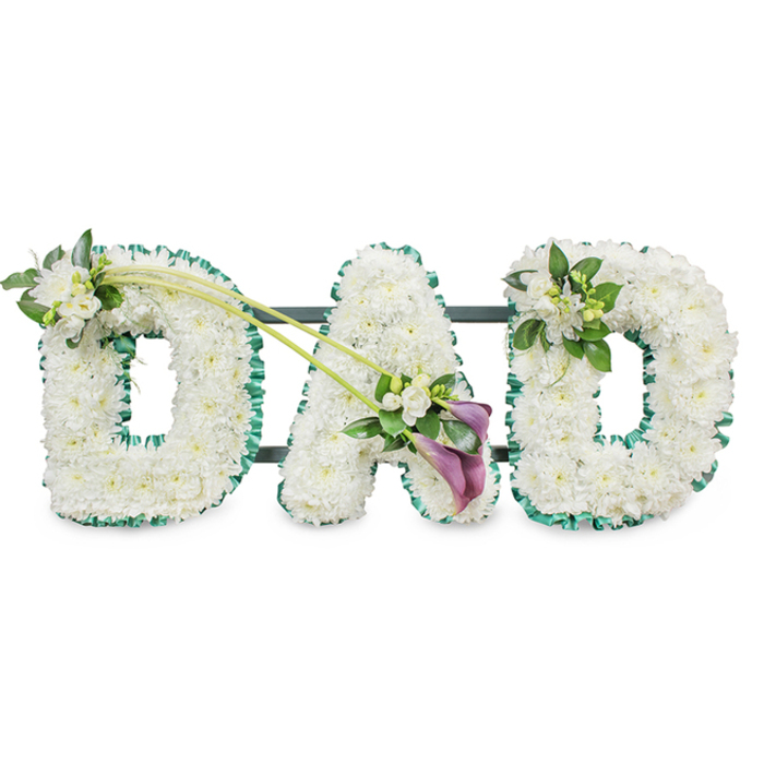 A solemn, meticulously assembled sympathy tribute forming the word DAD in large, three-dimensional letters-each letter composed of hundreds of small, immaculate white chrysanthemums or similar button blooms, tightly set to create a plush, velvet-like surface. A rich emerald-green satin ribbon is painstakingly applied around the outer edge of each letter, the folded ribbon providing crisp definition and a soft reflective sheen that contrasts with the matte texture of the flowers. The arrangement is supported by a dark frame that shows as horizontal bars behind the letters, confirming its upright display format for a chapel, funeral hall, or graveside. Floral embellishments are placed with sensitivity: the first D is graced at its upper left with a cluster of fragrant white freesia and lush green foliage; two elegant long stems curve from the top of that D toward the central A, finishing in an artful spray where two deep purple calla lilies stand amid more white freesia and leaves, their trumpet shapes adding solemn beauty and depth. The final D carries a slightly smaller grouping of freesia and greenery at its upper right, creating visual harmony. The quietly scented freesia and the soft feel of the chrysanthemums invite reflection. As Flowers Golders Green we prepare tributes like this daily for families attending services at Golders Green Crematorium, local churches, or community halls near Golders Hill Park, and we can arrange respectful delivery across Golders Green-see flowersgoldersgreen.site for options and timings.