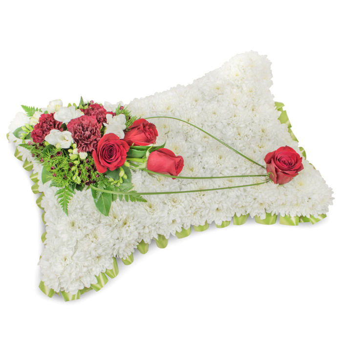A carefully composed memorial cushion, the rectangular pillow is entirely surfaced with dense, snowy white chrysanthemums whose tiny multi-petalled heads form a velvety carpet that speaks of peace and quiet rest. Arranged on the upper left is a striking spray of deep red roses and burgundy carnations - some tightly furled, others open - their rich colours set off by small clusters of white freesias and miniature spray chrysanthemums that provide delicate highlights. Lush greenery, from feathery fern fronds to more structured leaves, gives the arrangement a living framework, while three slender arcs of bright green bear-grass sweep diagonally across the white expanse to the lower right, guiding the eye to a single, perfectly shaped red rose that appears gently laid in place. The pillow is edged with a narrow band of light green satin ribbon that peeks out beneath the flowers, offering a neat, gentle frame to the composition. As an experienced local florist in Golders Green I craft pieces like this with sensitivity to the neighbourhood's many memorial services - from small chapel gatherings to tributes at Golders Hill Park remembrance spots - and Flowers Golders Green prepares each cushion to balance visual calm, subtle scent, and thoughtful symbolism. The overall impression is one of reverence and lasting affection: quiet, structured, and crafted to comfort those who come to remember.