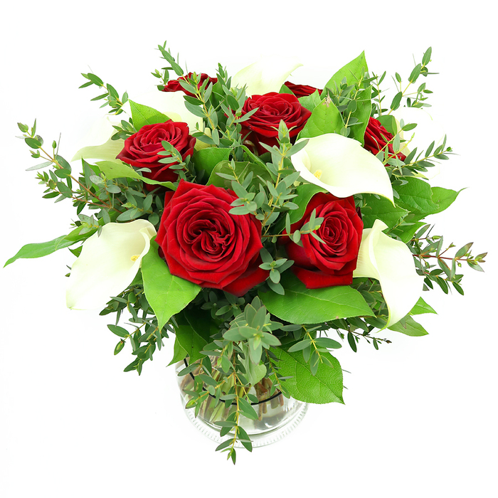 This photograph shows a sophisticated floral arrangement of deep red roses and snowy white calla lilies, gathered into a rounded, balanced bouquet and displayed in a transparent glass vase on a stark white background. At the core of the design, several fully opened red roses unfurl layer upon layer of velvety petals, each blossom capturing a saturated crimson hue that suggests romance and enduring affection. Interspersed among them, graceful calla lilies lift their elegant, trumpet-shaped forms, the smooth, ivory petals curving in clean lines that reveal a subtle yellow-orange spadix at the centre of each bloom. The juxtaposition of the rich red roses and the refined white callas creates a dramatic yet tasteful contrast, reminiscent of a polished dinner setting in a Golders Green home near Brent Cross or the leafy avenues of Hampstead Garden Suburb. Surrounding the focal flowers, an abundance of green foliage adds depth and movement: slender, slightly drooping eucalyptus stems weave through the design, their soft, silvery-blue leaves lending a gentle, airy feel, while larger, glossy green leaves form a solid backdrop that intensifies the vibrancy of the red and white blooms. The stems are neatly aligned inside a simple cylindrical vase with delicate ribbing toward the base, and the clear water inside hints at the freshness of flowers prepared the same day by a local Golders Green florist. Bright, even lighting floods the arrangement, allowing every texture-from the waxy sheen of the calla lilies to the feathered edges of the roses-to be clearly seen. The overall impression is one of refined celebration, ideal for anniversaries, romantic gestures, or as an elegant centrepiece for gatherings at home or intimate dinners in the local area.