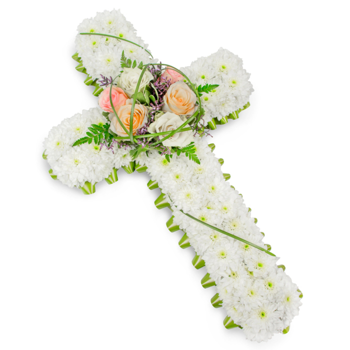 This image captures a traditional Christian cross rendered entirely in flowers, arranged with the kind of care and precision you might expect from an experienced Golders Green florist preparing a final tribute for a local family. The body of the cross is densely carpeted with small, neat white chrysanthemum heads, placed so closely together that they create a seamless, plush surface. Their uniform, rounded shapes and crisp white petals give the cross a soft yet structured look, suggesting purity and a sense of calm. Along the perimeter of the cross, from the top arm down to the base, a slim line of vivid green foliage and supportive floral materials frames the white chrysanthemums. This green edging subtly accentuates the contours of the cross, adding both contrast and a touch of natural freshness that recalls the nearby green spaces of Golders Hill Park or Hampstead Heath. At the central intersection, a graceful cluster of larger flowers forms a raised focal point. Here, several roses in layered shades of peach, coral-pink, and creamy ivory gently open, their velvety petals overlapping to create a soft focal glow. Threads of bright green grass loop and weave among the rose heads, while delicate fern fronds and wisps of small purple flowers add light, feathery texture around the edges of the spray. The lighting is even and bright, emphasizing the clean whites and vivid greens without harsh shadows. This understated yet heartfelt funeral cross feels ideal for a church or chapel service near Golders Green, a memorial gathering at a local family home, or a graveside farewell, offering a meaningful symbol of faith, remembrance, and abiding love.