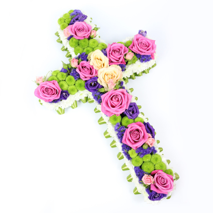 This image shows a traditional floral cross displayed on a brilliant white background, the camera positioned slightly above to capture the full shape and intricate detail of the design. The cross is elegantly defined by a border of tightly clustered white spray chrysanthemums, creating a clean, cloud-soft frame that suggests purity and peace. Nestled just beneath that border, a ribbon of folded light green material-possibly satin ribbon or carefully arranged foliage-traces the edges, lending a fresh, modern note reminiscent of the leafy avenues around Golders Green Road. Inside this refined outline, the heart of the cross is a vivid mosaic of colour and texture. Bold hot pink roses and softer, creamy peach-to-pale-yellow roses are arranged in generous clusters along the length and arms, each bloom fully open to reveal its spiralling petals. Between these larger focal flowers, small, rounded lime-green button chrysanthemums dot the surface like cheerful gemstones, their compact, textured heads bringing energy and a sense of new growth. Interwoven among the roses and green mums are smaller blooms in rich purple and deep violet tones-possibly lisianthus or statice-adding depth, contrast, and a quietly reverent mood, suitable for a funeral service or memorial gathering in Golders Green. The composition is full and meticulously balanced, with no gaps, so the flowers appear to form a continuous, living tapestry. Bright, even lighting ensures every nuance of petal, leaf, and ribbon is clearly visible, highlighting the craftsmanship of an experienced local florist. This floral cross feels like a heartfelt expression of sympathy and faith, an uplifting tribute that honours a life while offering comfort to grieving family and friends.