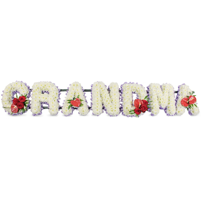 A lovingly crafted floral memorial displays the word GRANDMA in strong, upright block letters, arranged in a straight horizontal line as if ready to rest at the front of a service in Golders Green. Each character is made from tightly clustered white cushion chrysanthemums, their small, rounded petals forming a soft, pillowy surface that appears both full and gentle to the touch. Along the edges of each letter runs a narrow trim of pale purple ribbon or backing, adding a delicate outline that hints at lilac tones and provides subtle contrast against the pure white blooms. Carefully placed red accents bring warmth and emotion to the tribute. At the start, the letter G is embellished with a rich spray of deep red roses nestled into its curve, alongside a distinctive red-and-white variegated calla lily that draws the eye and gives the design a unique signature. The first A carries a single heart-shaped red anthurium positioned near its base, the glossy, sculptural bloom creating a beautiful counterpoint to the soft chrysanthemum texture. In the centre, the letter M holds another cluster of red roses paired with a second red anthurium, while the final A is finished with a solitary anthurium bloom that balances the composition. The letters sit on a discreet dark green metal frame with hints of foliage, providing stability while remaining visually unobtrusive. Crafted by an experienced local florist familiar with the gentle, family-focused character of Golders Green, this bespoke funeral flower arrangement offers a deeply personal way to honour a grandmother's life, suitable for church, crematorium, or graveside tributes across the area.