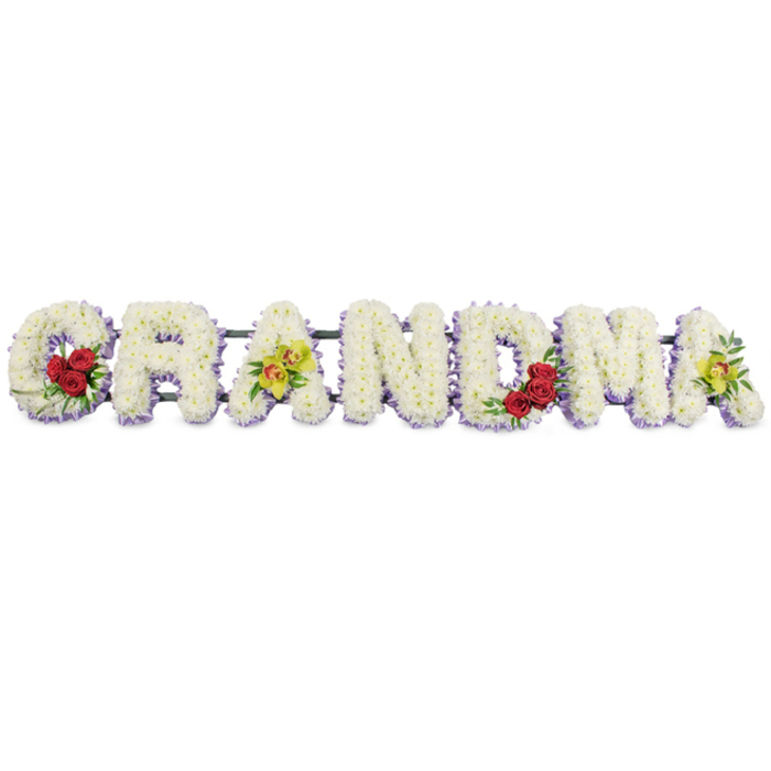 A touching floral display spells out GRANDMA in large, ornate block letters, arranged in a straight line as if ready to rest beside a coffin or at the front of a chapel in Golders Green. The letters are crafted from a dense tapestry of small, snow-white chrysanthemum heads, each bloom carefully wired and nestled so that no gaps show, giving the whole word a soft, cushioned look. Underneath this white blanket, a slim band of pale lavender or light purple flowers peeks out around the base of each letter, creating a gentle outline that adds depth without overpowering the purity of the white. On the sweeping curve of the initial 'G', three velvety red roses sit low to the side, framed by deep green leaves that bring a natural richness and a sense of heartfelt love. The first 'A' from the left features two yellow-green cymbidium orchids, their graceful petals and speckled reddish-brown centres set carefully on the crossbar, lending an almost heirloom elegance. The letters 'R', 'N', and 'D' are left entirely in white and lavender, their simplicity allowing the tribute to breathe and convey quiet respect. The strong, central arches of the 'M' cradle a second little cluster of red roses to the lower right, visually echoing the first accent on the 'G'. At the far end, the final 'A' repeats the yellow-green orchids, so the design feels balanced from start to finish. A slender dark green or black metal frame connects all the letters subtly along the top, making the piece sturdy enough for transport to local crematoriums, synagogues, or cemeteries. This elegant sympathy arrangement offers a deeply personal farewell to a cherished grandmother, reflecting both purity and enduring love.