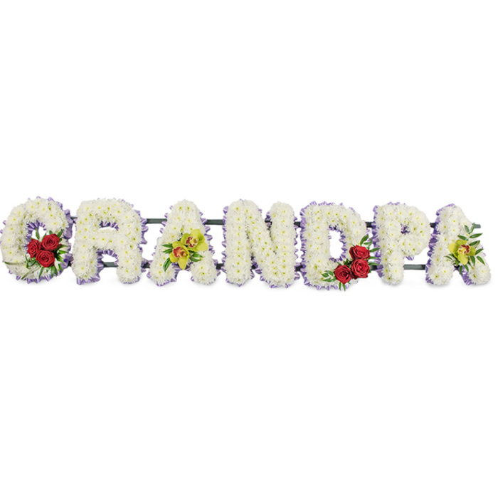 A refined floral memorial spelling GRANDPA stretches horizontally across a pure white background, every letter formed from masses of white chrysanthemums that create a soft, cushioned effect, like a quilt of petals laid in tribute. The letters are generously proportioned and clearly legible, their strong block shapes softened by the tiny, rounded chrysanthemum heads. Around each letter runs a narrow band of lavender-purple ribbon, just visible at the edge of the blooms, offering a gentle halo of colour that subtly defines the silhouette of the word. In the curve of the opening G, three to four deep red roses are arranged in a tight cluster, their velvety petals unfurling against the white, accompanied by small sprigs of glossy greenery that add texture and depth. The third letter, A, stands out with a graceful spray of yellow-green orchids, some with fine reddish markings at their centres, lending an elegant and slightly exotic note. This balance of accents is continued as the rounded centre of the D features another cluster of red roses, mirroring the G, while the final A is again adorned with orchids, completing a thoughtful rhythm of colour along the tribute. Discreet horizontal bars and a dark frame behind the flowers support the structure, keeping the letters perfectly aligned while remaining largely hidden from view. The arrangement feels calm and respectful, ideal for a funeral or memorial service in Golders Green, whether placed at the crematorium chapel or in a local synagogue. With its serene whites, passionate reds, and graceful orchids, the piece carries a message of purity, love, and dignified remembrance for a cherished grandfather.