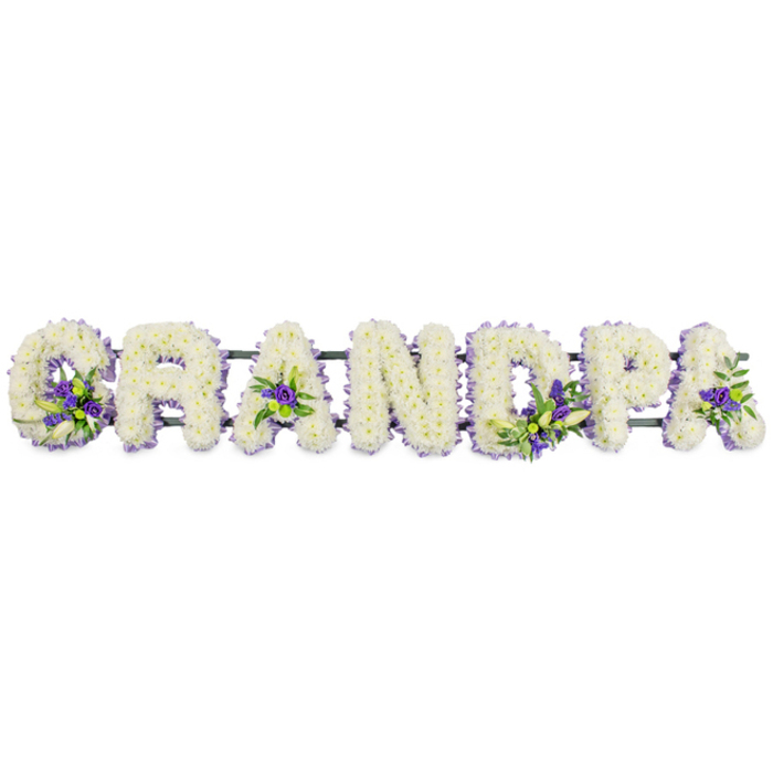 The image shows an elegant funeral letter tribute spelling out the word GRANDPA in large, separate block letters that sit side by side in a clear horizontal line, reminiscent of a display at a memorial gathering in the Golders Green area. Each letter has been meticulously built up with countless small white chrysanthemum or carnation blooms, their petals overlapping in tight clusters so that the surface appears plush and even, like a white quilt of flowers. The hue is a calm, creamy white rather than stark, gently diffusing the light and giving the letters a soft, reverent glow that feels appropriate for a final farewell. Interspersed through this sea of white are carefully positioned clusters of purple accent flowers, perhaps small irises, hyacinths, or lisianthus, cradled by sprigs of fresh green foliage. These accents create moments of colour at key points: within the round of the 'G', sitting in the open triangle of the first 'A', accenting the curve of the 'D', and adding life to the upright stem of the 'P' and the last 'A'. The greens and purples bring depth and gentle contrast, much like pockets of colour among the trees and paths around nearby Golders Hill Park. A dark green support bar runs just underneath the letters, mostly hidden, linking the tribute into one cohesive piece that can be easily placed near a coffin, on a hearse, or beside a photo of the grandfather being remembered. This floral tribute feels deeply considered, a personalised expression of love and respect that a local Golders Green florist might create for families saying goodbye to a cherished grandparent.