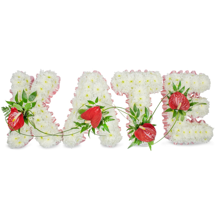 An elegant, personalised floral name tribute spells KATE in large capital letters, each letter built up from a dense, uniform carpet of tiny white chrysanthemums that create a soft, cloud-like texture across every face and curve. Interspersed among that pristine white are vivid red anthuriums with their glossy, heart-shaped spathes and upright, pale spadices; these bold red forms are placed thoughtfully to provide warm focal points and to catch the eye against the white field. Dark green foliage - from fern fronds to broad glossy leaves - frames the anthuriums and is woven through the white blooms, adding depth, structure and a natural cadence to the lines of each letter. A pale pink ribbon or pleated trim follows the outer edges of the letters, offering a gentle contour and a modest, refined colour accent that enhances rather than distracts. Fine green vines drape and bridge between the letters, suggesting continuity and unity while a soft shadow beneath the composition lifts the letters away from the bright white background, creating a subtle three-dimensional effect. The overall mood is one of quiet dignity and careful craft, a bespoke funeral or memorial piece suitable for services across Golders Green - from a chapel near Finchley Road to a family home close to Temple Fortune - where families often look for tributes that combine classic sympathy flowers with contemporary, heartfelt design.