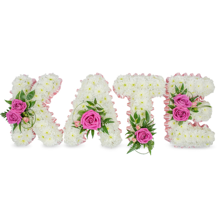 A bespoke sympathy tribute meticulously spells KATE in bold, three-dimensional letters, each letter painstakingly constructed from hundreds of small, pristine white chrysanthemums that create a seamless, cloud-like surface. The inner contours are gently traced with a pale pink satin ribbon, a delicate flourish that softens the architectural block letters and directs the eye toward the floral focal points. Vivid hot pink roses-bright, saturated fuchsia-are placed with thoughtful balance: two open roses on the K's left arm framed by mixed greens and spiral grass, a single prominent rose nestled low within the A, a solitary rose anchoring the base of the T beside distinctive fern-like fronds, and two roses clustered along the right side of the E highlighted by variegated leaves and tiny pink accents. Subtle sprays of light pink alstroemeria and small spray roses mingle with glossy ruscus and eucalyptus hints, offering a modern, garden-fresh edge to the composition. The chrysanthemums are densely packed for a plush, tactile finish while the satin ribbon and contrasting foliage sharpen the letters' silhouettes; soft studio lighting and the faint shadows beneath each letter reinforce the arrangement's freestanding, three-dimensional character. As prepared by Flowers Golders Green for families in the local area, this arrangement is well suited to services at Golders Green Crematorium or a celebration of life at a nearby chapel or community hall, communicating dignity, tenderness and a personalised sense of farewell from an experienced neighbourhood florist.