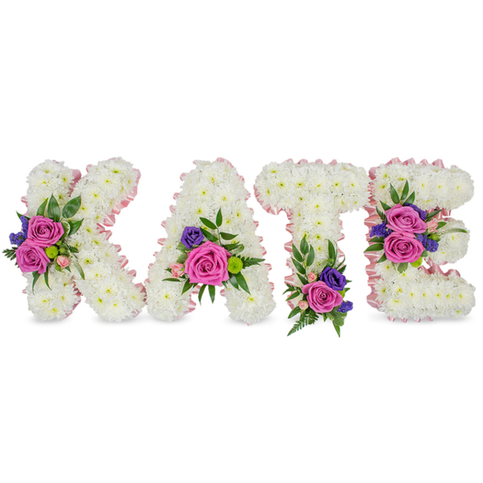 A carefully crafted standing tribute spells the name KATE in four distinct, three-dimensional letters constructed from thousands of tight, snowy-white chrysanthemum heads. The chrysanthemums form a continuous, dense surface whose small, ruffled petals create a uniform, plush texture across each character. Each letter's silhouette is subtly emphasized with a pale pink satin ribbon that follows both outer and inner edges, introducing a gentle warmth against the immaculate white. Across the letters sit intentionally placed accent clusters: prominent fuchsia-pink roses - their velvety petals rounded and full - are paired with smaller pastel pink spray roses and interspersed with fine deep purple or blue blooms, probably statice or diminutive lisianthus, to add tonal depth and a hint of dusk. Greenery, from lacy fern fronds to smooth eucalyptus leaves, frames and supports these colour groups, lending vertical lines and soft movement that prevent the design from feeling static. Compositionally, the K features a bold diagonal cluster, the A is grounded by a floral grouping at its lower right curve, the T carries a base cluster to anchor it, and the E is softened with two placements on its middle and lower bars - a considered distribution that balances the name visually. Photographed against a bright white backdrop, this piece reads as a compassionate, personalised sympathy tribute suitable for funerals, memorial services, or graveside ceremonies in Golders Green, often requested by families attending services near Hoop Lane or the local crematorium. The overall effect is tender, precise, and quietly beautiful.