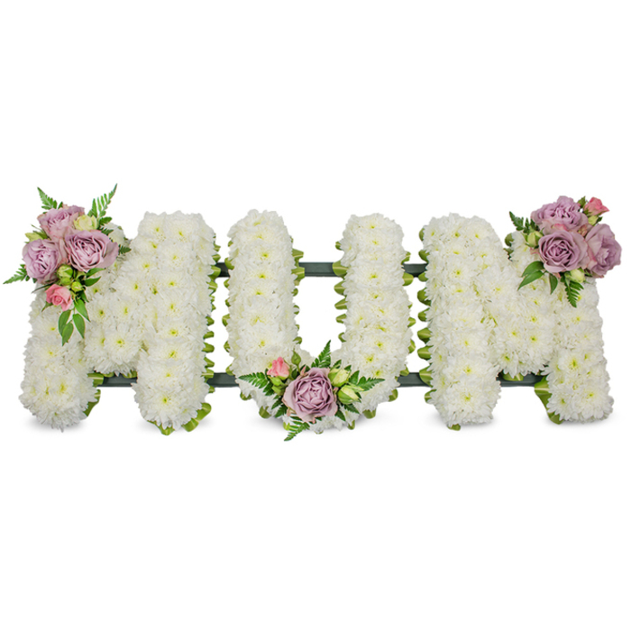 A lovingly made, large standing tribute spells out MUM in capitally formed letters, each letter a dense carpet of small, bright-white chrysanthemums or closely packed white spray flowers that create a cloud-soft texture and a pristine, even surface. The composition sits against a pure white backdrop under soft, diffuse lighting, which highlights the slight sheen on chrysanthemum petals and the tiny shadows that give the letters a sculpted, three-dimensional presence. To bring gentle colour and human warmth, three small clusters of muted lavender roses and pale pink buds are carefully placed: one on the upper left arm of the first 'M', another nestled within the central dip of the 'U', and the third adorning the upper right of the final 'M'. These roses, some in full bloom and some as delicate buds, offer a velvety contrast both visually and in scent, their faint perfume mingling with the crisp green aromas of fern fronds and glossy foliage woven into the design. A dark green supporting frame - likely ribbon-wrapped or mossed timber - runs subtly beneath the letters, visible as a darker contour that secures the shape while remaining unobtrusive. This classic sympathy arrangement expresses respect, remembrance, and tender love for a mother, and is the sort of piece we prepare with care at Flowers Golders Green for services near Golders Green Crematorium, local chapels, or gravesides across the area from Hendon to Hampstead Garden Suburb. The overall mood is one of calm dignity, crafted to offer comfort and quiet reflection.
