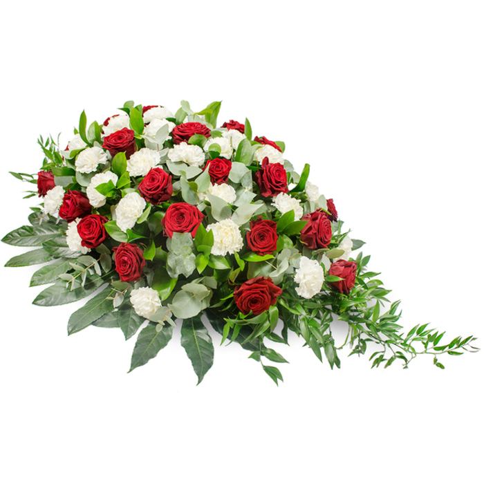 A refined funeral floral spray is shown in full detail against a white studio background, allowing its shape and colours to be clearly appreciated. The arrangement is composed in a low, elongated mound, with a generous, rounded fullness to the left that gradually narrows into a tapering, trailing end on the right. Deep red roses, each bloom perfectly shaped with layers of velvety petals, rise slightly above the rest of the flowers, lending height and focus to the tribute. Between and around these roses are plentiful white carnations, their ruffled, intricate petals appearing almost like soft fabric, offering a gentle texture and a sense of purity and remembrance. The classic pairing of dark crimson and crisp white creates a solemn yet comforting palette, often chosen for funeral services and memorials. Around the blooms, a bed of verdant foliage completes the design. Broad, dark green leaves provide structure and give the spray its defined outline, while softer, silvery-green eucalyptus leaves weave through the roses and carnations, adding hints of muted colour and a calm, aromatic note. At the right-hand edge, lighter green, feathery stems arc outward in a natural cascade, softening the silhouette and suggesting quiet movement. The overall composition feels thoughtful and balanced, neither overly formal nor casual, ideal for honouring someone who was deeply loved yet appreciated for their simplicity. This is the sort of memorial piece that could be placed in a chapel of rest or delivered to a funeral home serving families in Golders Green, reflecting the area's sense of community and offering a tender, respectful farewell.