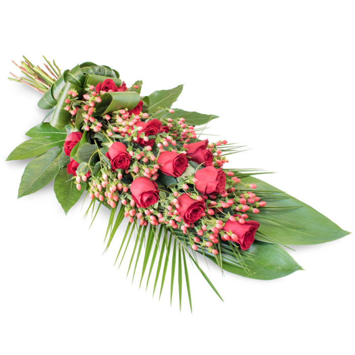 Set against a pristine white background, this long, graceful funeral spray of red roses and berries appears almost to float, every stem and petal clearly defined. The arrangement starts at the upper left where the stems are gathered into a tight bundle, neatly encased in a broad band of wrapped green leaf that gives a tailored, professional finish often seen in high-end tribute work around Golders Green. From that focal point, the spray extends diagonally downward, forming an elongated teardrop or soft crescent shape. Along its spine, deep, velvety red roses are meticulously spaced, some still in bud, others just unfurling, and a few fully open with layered petals that speak of deep love and respect. Interwoven through the roses are plentiful clusters of coral-pink to pinkish-red Hypericum berries, adding gentle volume and a slightly lighter, more playful red tone that contrasts with the richness of the petals. Supporting this central display is a foundation of lush greenery: broad, dark green leaves with a glossy sheen, reminiscent of Salal or Ruscus, create a solid base, while slender palm fronds and airy foliage sweep outward, particularly along the lower right edge, giving the spray movement and an elegant trailing effect. Fine, linear stems and smaller leaves add subtle complexity, catching imaginary highlights like dappled light might in one of the tree-lined streets near Golders Hill Park. The overall feel is dignified, serene, and carefully composed - a traditional sympathy tribute ideal for a funeral, cremation, or memorial gathering in and around Golders Green, conveying heartfelt condolences and the quiet beauty of a final farewell.