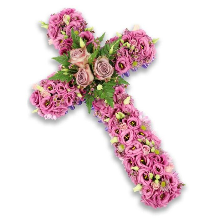 A refined funeral cross floral arrangement stands out sharply against a clean white background, its three-dimensional structure completely cocooned in blooms so that the visual focus is all on colour and texture. The palette is a gentle blend of soft pinks, mauves, and lilacs, creating an atmosphere of calm and tenderness. At the point where the arms intersect, three large roses in a dusty-rose and lavender-pink mix are in full bloom, their curling petals forming gentle rosettes that seem to open towards the viewer. These are surrounded by a halo of finely cut green fern fronds and shorter, rounded leaves, which help to frame the roses and add depth. From this central grouping, the stems and arms of the cross are densely layered with lisianthus and trumpet-shaped blossoms in a spectrum of pinks, some petals marked with darker centres that give a sense of quiet intensity. Nestled among them are tiny pink and lilac blooms, crisp white accents, and fresh light green buds that keep the arrangement from feeling heavy, suggesting renewal even within a tribute of farewell. Along the outer edges of the cross, thin slices of deeper purple ribbon or backing material peek out slightly, lending a subtle, tailored border. The overall impression is one of thoughtful craftsmanship and compassionate intent, the kind of floral cross that could rest gracefully in a chapel near Golders Green Crematorium or at a graveside in Hoop Lane Cemetery, offering a visual prayer of peace, remembrance, and gratitude for a life well lived.