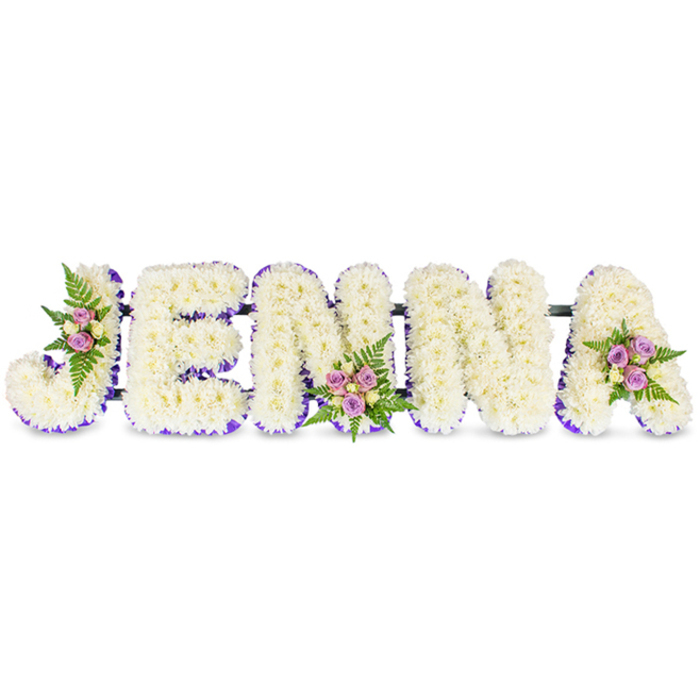 A bespoke, deeply personal tribute spells JENNA in bold, three-dimensional letters, each surface densely carpeted with hundreds of small creamy-white chrysanthemums that give the wording a soft, cloud-like relief against a spotless white background. A narrow, vivid purple trim outlines the inner edges and bases of the letters, providing crisp definition and an understated sense of dignity. Three carefully placed clusters - one on the J's curve, another on the lower centre of the second N, and a final grouping on the upper right of the A - introduce blush-lilac spray roses, a few pale cream roselets and sprigs of rich green fern, offering warmth, scent and natural contrast amidst the sea of white. Thin, dark metal bars link the letters so the tribute reads as a single, stable unit suitable for standing displays at a chapel or near a hearse. The arrangement is photographed in soft, diffused light that brings out the chrysanthemums' fine textures and the satin sheen of the purple ribbon, suggesting both comfort and quiet dignity. As a Golders Green florist accustomed to commissions for local services - from the crematorium on Hoop Lane to small family wakes near Golders Green Road and the community around the tube - Flowers Golders Green creates tributes that balance readability from a distance with intimate floral detail up close. This name tribute offers families a crafted, heartfelt expression of remembrance, appropriate for funeral settings, memorial tables, or a chapel doorway where the name itself becomes the focal point of love and farewell.