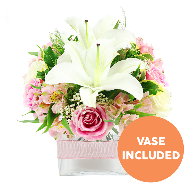 Against a clean, uncluttered white background, a lush floral arrangement in a clear square glass vase draws attention with its soft whites, pinks, and fresh greens, creating a tranquil yet joyful mood. Two fully open white Oriental lilies stand proudly in the centre, their broad petals arching outward like small trumpets and their rich reddish-brown stamens and pistils clearly visible, adding depth and a hint of drama. Encircling these lilies is a ring of pink roses in various stages of bloom: tight buds with spiralled petals suggesting new beginnings sit beside fully open roses whose ruffled edges and generous heads feel luxurious, evoking classic romantic gestures so often requested by Golders Green customers for anniversaries and special milestones. Threaded throughout are dainty stems of alstroemeria in shades of light pink and creamy white, many with subtle yellow and darker speckling inside their petals, lending the bouquet a painterly, intricate look. Fine clusters of white baby's breath nestle between the main flowers like tiny clouds, softening transitions and filling small gaps so the arrangement feels complete from every angle. Surrounding all this, a variety of textured foliage-broad, deep green leaves polished to a gentle shine, along with lighter, feathery greens-creates a natural frame that enhances both colour and shape, reminiscent of the greenery along nearby Hampstead Garden Suburb. The stems are gathered closely within the low, square vase, their cut ends visible through the clear glass, giving a sense of honest freshness. Around the midpoint of the vase, a light pink ribbon is wrapped horizontally, offering a hint of decorative detail that suggests the bouquet is ready to present as a thoughtful gift for birthdays, thank-yous, or a comforting delivery to a home near Golders Green station. In the bottom right of the image, a bright orange circular label with strong white capital letters clearly reads VASE INCLUDED, indicating that the glass container comes with the arrangement.