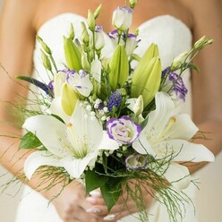 Cradled in gentle hands and set against the understated elegance of a white strapless wedding dress, this bridal bouquet finds its own poetry amidst the leafy avenues of Golders Green. Large, pristine Oriental lilies bloom at the center, their petals creamy-white, each highlighted by textured dots and revealing vibrant yellow stamens-unmistakable signatures of purity held dear throughout local wedding traditions. Tracing around them, lisianthus buds unfold into ruffled elegance, their white petals tinged at the edges with just a kiss of royal purple. Tucked between are clusters of deep violet veronica, adding depth and intensity like the changing light across Hampstead Garden Suburb. Airy sprigs of baby's breath drift throughout the bouquet, their miniature blooms like a soft morning mist, softening every line and contour. The rich mix of free-flowing greens-broad leaves, unopened lily buds, and feathery asparagus fern-spill gently, echoing the wild charm of Golders Hill's hidden gardens. The bouquet, balanced in form yet brimming with natural energy, is a tribute to both tradition and the fresh romance of a wedding day. Each flower's fragrance mingles tenderly, a reminder of spring's arrival on Golders Green Road, made for the bride who seeks not just beauty but authentic connection to her local story.