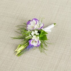 A delicately styled buttonhole rests on a pale beige, linen-like fabric, the gentle weave and soft light creating an almost hushed backdrop, as if the room is still before guests arrive. At the centre, two white lisianthus blossoms unfurl in layered, ruffled petals, each one tipped in a rich, vibrant purple that deepens along the edges like ink brushed on fine paper. They are accompanied by airy clusters of tiny white filler blooms resembling baby's breath, which bring a sense of lightness and space, much like the open sky above Golders Green's tree-lined avenues on a clear day. Rising beside them, a single upright stem of deep purple muscari, its tight bell-shaped flowers stacked closely together, adds a quietly dramatic texture, subtle yet distinctive. Surrounding this focal trio is a nest of mixed greenery: slender, feathery fern-like fronds and a few slightly broader leaves that suggest freshness and the faint scent of cut stems, as though gathered at first light. Two small, pale green buds, still firmly closed, hint at future blossoms and the promise wrapped up in the occasion. At the base, the stems are neatly aligned and wrapped with a smooth, white satin ribbon, carefully secured to form a slim, elegant handle ready for pinning. The ribbon's cool sheen contrasts gently with the organic forms above, balancing wildness with formality. The entire boutonniere feels composed for a meaningful moment-a groom stepping out of a townhouse just off Finchley Road, a prom-goer meeting friends near Golders Green station-carrying with them a quiet, floral whisper of the day's importance.