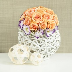 A sophisticated floral centerpiece is arranged with the quiet confidence of an experienced North London florist, poised on a smooth white surface against a softly textured beige background that recalls the calm interiors of homes around Golders Green and Temple Fortune. The focus of the piece is a lush, spherical cluster of peach and apricot roses, each bloom carefully positioned so that their velvety petals overlap seamlessly, creating a smooth, domed silhouette. The roses glow with warm pastel tones, from soft blush to richer apricot, capturing that gentle light you often see on early evenings near Golders Hill Park. Encircling the base of this rose globe is a delicate band of white lisianthus, each flower rimmed with a rich purple edge that reads almost like watercolour paint along the petal tips. Interspersed among them are fine stems of white baby's breath, adding tiny constellations of blossoms that soften the transition between the roses and their collar of colour. The flowers are gathered into a white woven vessel, broadly rounded and slightly bulbous, created from interlacing strands of wicker or rattan. Its open, airy lattice allows glimpses of shadow and negative space, lending the arrangement a contemporary feel with a hint of rustic charm that would suit both modern Golders Green apartments and traditional houses near Hampstead Garden Suburb. In the foreground, two decorative spheres rest on the same white surface, woven from pale, natural-toned wood strips that echo the weave of the main container while adding playful variation in size and form. Everything is framed by the warm neutral fabric backdrop, which enhances the peach, white, and purple tones without distraction. The overall effect is one of poised romance and understated luxury, perfect for wedding table settings, anniversary dinners, or stylish celebration lunches hosted locally.