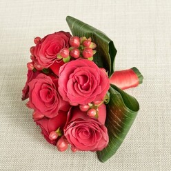 A beautifully composed bouquet of deep red roses and berries lies on a softly textured, light beige fabric, evoking the calm, understated elegance of a townhouse interior just off Golders Green Road. The arrangement is compact yet full, with around six large red roses forming the main focus, their velvety petals catching the light in rich tones of crimson and garnet. The roses are in perfect bloom, their centres still gently furled while the outer petals curve outward in smooth, spiralling layers that invite you to imagine their cool, satiny feel. Scattered among the roses are clusters of small, round Hypericum berries in shades of red and flushed orange, each berry held on a slender green stem that adds a fine, intricate texture between the larger blooms. Framing the bouquet are several glossy, deep green leaves, broad and tropical in shape, that have been expertly curled and wrapped around the flowers to create a neat, architectural collar, giving the design both structure and drama. A narrow band of matching red satin ribbon encircles the stems, partly visible at one side, its sheen echoing the depth of the petals and providing a polished finishing touch. The bouquet is photographed from a slightly elevated angle, with soft, even lighting that highlights every contour without overwhelming the natural colours. This design feels perfect for a bridesmaid's bouquet, an engagement photo shoot near Golders Hill Park, or a romantic same-day delivery across Golders Green, capturing that balance of passion and refinement that makes classic red roses so timeless.