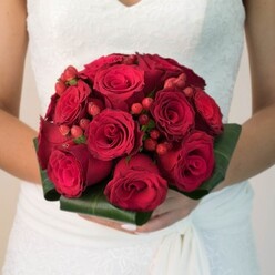 This evocative photograph, reminiscent of a cherished moment along Finchley Road, places a lush, handcrafted bridal bouquet at its centre, held tenderly by a bride whose hands and gown reflect Golders Green sophistication. The arrangement is an artful display of velvety red roses, each flower's petals either tightly furled or softly unfolding, painting a picture of passionate elegance. Vibrant clusters of glossy red hypericum berries interlace with the blooms, bringing a subtle, festive sparkle that feels like a nod to the area's vibrant culture and markets. Encircling the bouquet, broad green leaves are folded into a flawless, luxurious collar, amplifying the drama of the reds while grounding the composition in natural beauty. Gentle light filters across the bouquet, accentuating every texture-the smooth petals, the plump berries, and the sturdy green foliage. The bride's wedding dress, with its delicate lacework, hints at tradition and contemporary charm, much like the classic houses of Golders Green. This arrangement, available from Flowers Golders Green, is the embodiment of romance and celebration, carrying a sense of place and memory that would resonate with couples planning their special day in North West London.