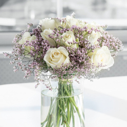 This image presents a thoughtfully composed sympathy bouquet, dominated by softly glowing white roses and airy purple filler flowers, arranged in a tall clear glass vase that stands on a bright, uncluttered surface. Each rose is fully opened, its layers of creamy, almost porcelain-like petals unfurling in gentle spirals that suggest both purity and quiet strength. Around and between the roses, fine sprays of lavender and light mauve statice create a misty border, their small, cloud-like blossoms bringing a dreamy, almost weightless texture to the design. The green stems rise in an orderly cluster from the base of the bouquet, then continue straight down into the water, clearly visible through the transparent cylinder and giving the composition a feeling of honesty and simplicity. The water itself looks freshly changed, crystal clear, as if the bouquet has just been prepared by an experienced florist familiar with the slower, reflective pace of Golders Green's residential streets and nearby parks. Soft, diffused daylight floods the scene from one side, catching the gentle curves of each petal and the subtle lilac tones of the filler flowers, while the background fades into an unobtrusive blur of pale grey and white. There are no distracting objects or patterns, only a sense of calm space around the arrangement. The overall impression is one of quiet solace and graceful respect, the sort of arrangement often chosen to send sympathy and support to families in Golders Green, Temple Fortune, and the surrounding neighbourhoods during times of loss and remembrance.