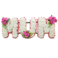 A deeply felt memorial arrangement forming the word MUM in three large standing letters, each letter composed of dense, pristine white chrysanthemums that create a uniform, velvety surface. The tiny, layered petals of the mums form a soft sculpted field, while a ruffled blush-pink trim hugs the outer edges of each letter, lending a gentle, comforting halo of colour. A dark green support frame peeks between the letters, giving the tribute its upright shape for display on a chapel easel or graveside plinth. Deliberate accent clusters bring moments of vibrant warmth: a small bouquet of fuchsia-pink roses with ferny greenery and coral hypericum berries nestles at the top-left of the first M; a matching posy sits in the base curve of the U; and the final M carries its coordinating spray on the top-right. The roses range from tight buds to half-open blooms, introducing varied texture and a deeper, rosy perfume that complements the clean, powdery scent of the chrysanthemums. Viewed under soft funeral lighting-perhaps in the chapel at Golders Green Crematorium or by a family home on Golders Green Road-the tribute reads as both dignified and tender. As designers at Flowers Golders Green we often choose this combination for mothers who cherished subtlety and grace: white for remembrance, pink for affectionate warmth, and the green structure for reassuring support. The piece is crafted to comfort, to be seen easily from a distance, and to provide a beautifully personal farewell.