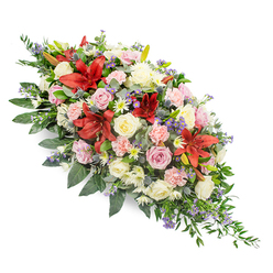 A large, double-ended funeral spray is displayed on a clean white background, its shape tapering smoothly towards each end, like a gentle exhale of colour and fragrance created for a final farewell in Golders Green. The design is dense and abundant, giving the impression of great care and intention in every stem placement. Deep red Oriental or stargazer-style lilies form the emotional heart of the piece, their velvety petals radiating strength and passion, with each bloom fully open to reveal pale centres and textured stamens. Around these dramatic focal flowers, clusters of creamy white and blush-pink roses are tucked closely together, some still in bud, others generously unfurled, suggesting a life remembered through many stages and cherished moments. Pale pink spray carnations fill the gaps, their frilled petals creating a soft cushion that contrasts beautifully with the bolder lilies. Small white daisy-like flowers and fine heads of lilac and lavender filler blooms add intricacy, like quiet whispers within the larger statement, with hints of purple and soft mauve threading through the design. Sprigs of blue-toned thistles and cool-toned fillers lend a subtle wildness and modern touch, echoing the natural textures found in nearby Hampstead Heath. The whole composition is supported by a rich collar of dark green foliage, including broad, glossy leaves and finer, trailing fronds that peek out at the edges, giving the spray a flowing, almost river-like outline. This arrangement feels deeply comforting and reverent, perfectly suited to accompany a coffin at a Golders Green funeral service or to be delivered as a heartfelt condolence tribute to a local family in mourning.